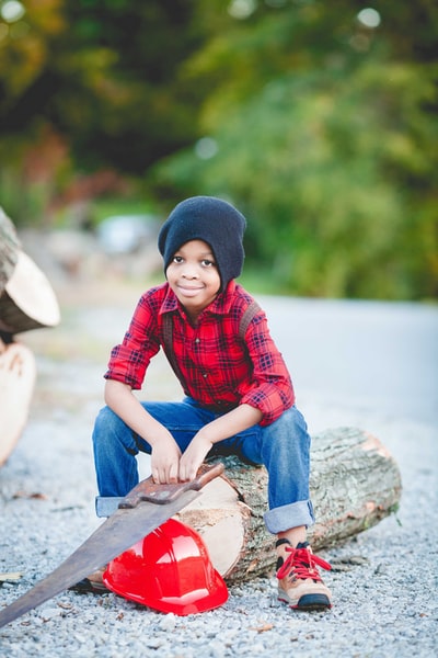 男孩坐在圆木上，手持手锯，戴着红色安全帽